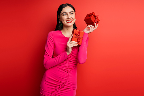 charming woman with brunette hair and trendy earrings smiling while standing in magenta party dress and holding wrapped gift boxes for holiday on red background