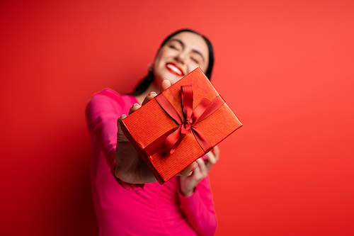 happy blurred woman with brunette hair smiling while standing in magenta party dress and holding wrapped present with ribbon for holiday on red background