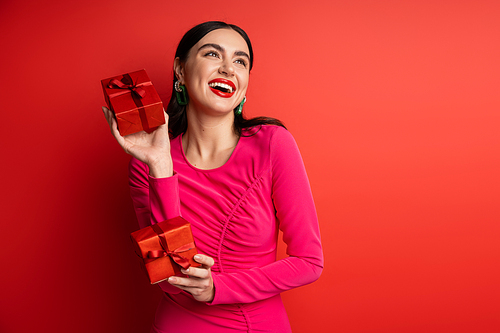 overjoyed woman with brunette hair and trendy earrings smiling while standing in magenta party dress and holding wrapped gift boxes for holiday on red background