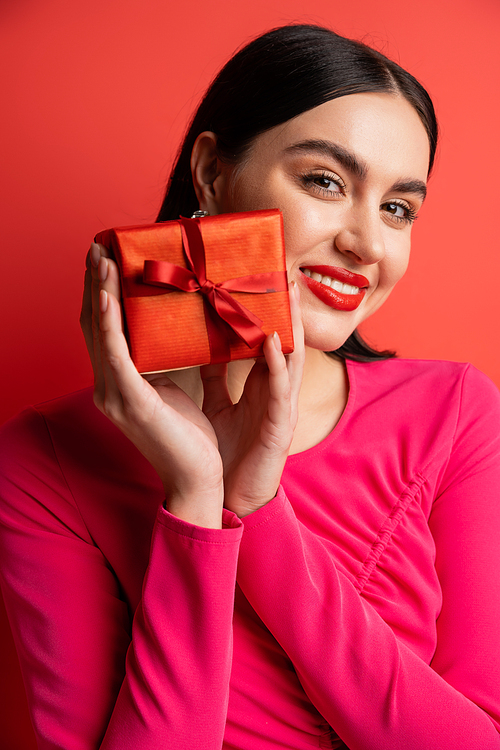 portrait of gorgeous woman with brunette hair smiling while standing in magenta party dress and holding wrapped surprise gift for holiday on red background