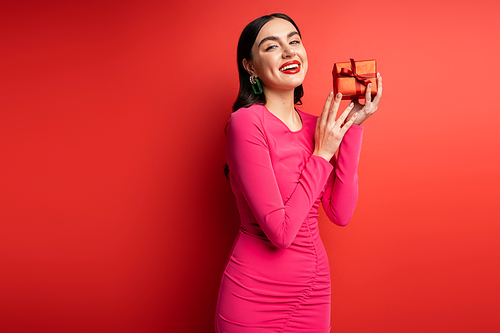 glamorous woman with brunette hair and trendy earrings smiling while standing in magenta party dress and holding wrapped surprise gift for holiday on red background