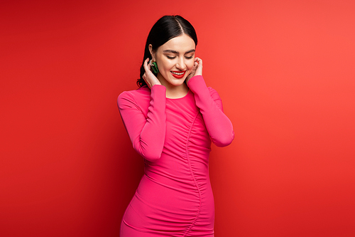 glamorous woman with brunette hair and trendy earrings smiling while standing in magenta party dress while posing and looking down on red background