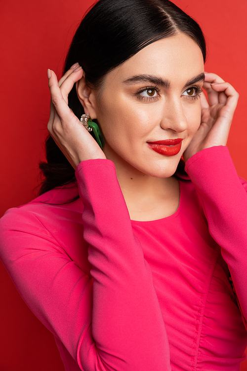 portrait of alluring woman with brunette hair and trendy earrings smiling while standing in magenta party dress while posing and looking away on red background