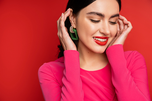 joyful and shy woman in trendy earrings adjusting her brunette hair and smiling while standing in magenta party dress while posing on red background