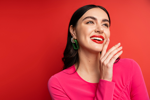 portrait of pretty woman with brunette hair and trendy earrings smiling while standing in magenta party dress while posing with hand near face on red background