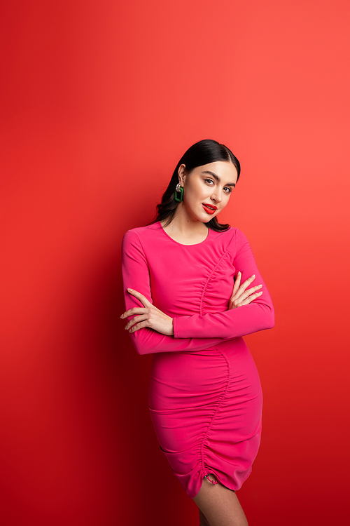 gorgeous woman with brunette hair and trendy earrings standing with folded arms in magenta party dress while posing and looking at camera on red background