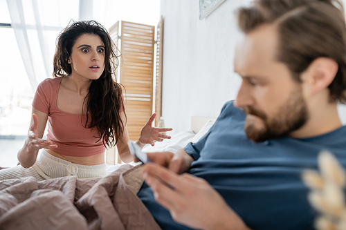 Annoyed woman in pajama quarrelling at blurred boyfriend with smartphone on bed