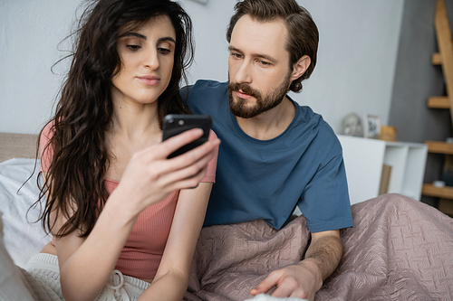 Brunette woman in pajama using smartphone near serious boyfriend on bed