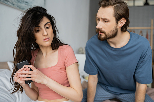 Brunette woman hiding cellphone near jealous boyfriend in bedroom