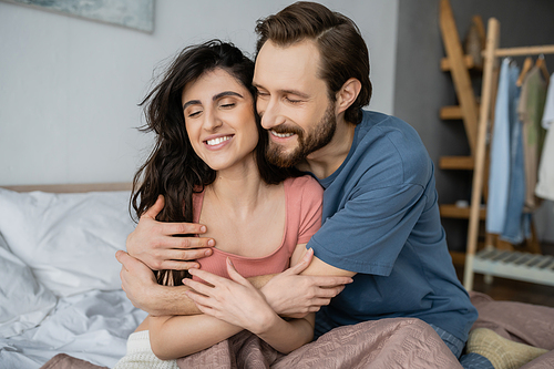 Overjoyed man hugging brunette girlfriend in pajama on bed at home