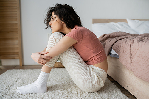 Side view of depressed woman in pajama sitting near bed at home