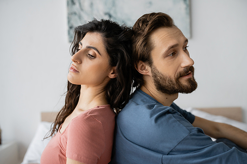 Sad couple sitting back to back in blurred bedroom at home