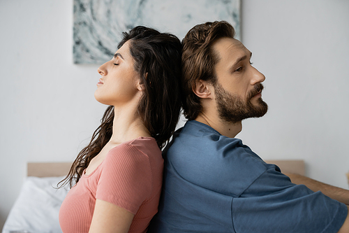 Upset bearded man sitting back to back with brunette girlfriend in bedroom