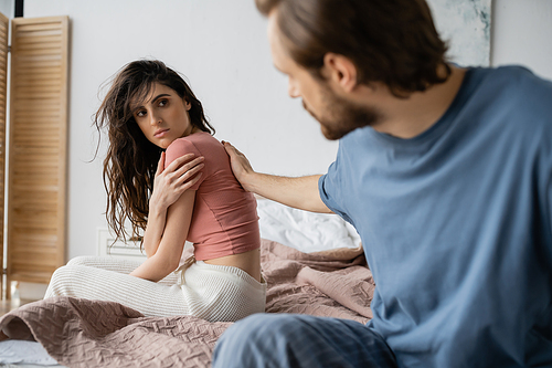 Blurred man touching offended girlfriend in pajama on bed at home