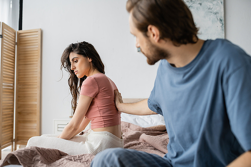 Blurred man touching disappointed girlified in pajama on bed