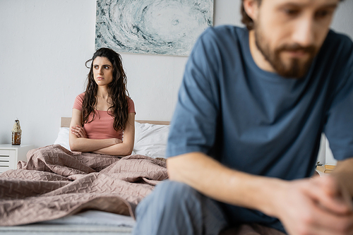 Angry brunette woman crossing arms while sitting near blurred boyfriend in bedroom