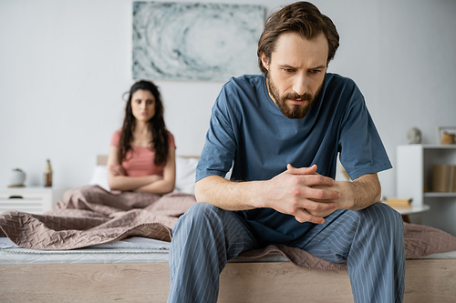 Tensed man in pajama sitting near blurred girlfriend in bedroom at home