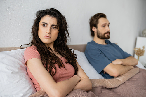 Nervous woman crossing arms while sitting near blurred boyfriend on bed