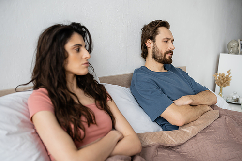 Dissatisfied man crossing arms while sitting near blurred girlfriend on bed at home