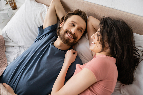 Top view of smiling brunette woman looking at bearded boyfriend on bed in morning