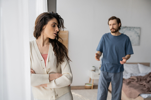 Displeased woman crossing arms near blurred boyfriend quarrelling at home