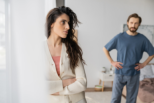 Angry woman crossing arms while standing near blurred boyfriend in bedroom