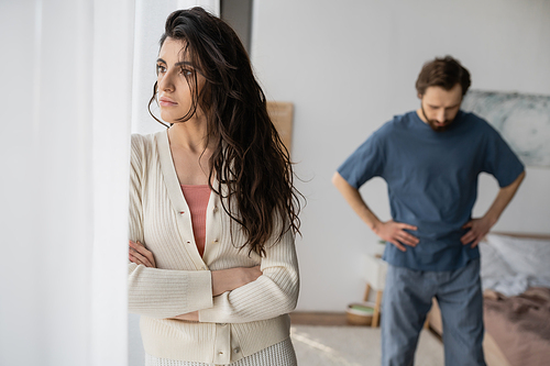 Displeased woman standing near curtains during relationship crisis with blurred boyfriend at home