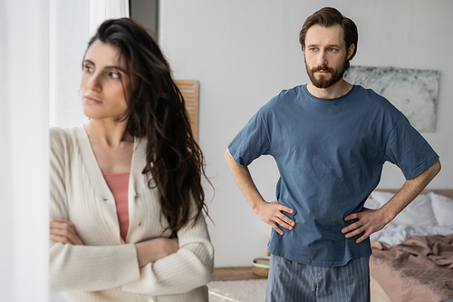 Bearded man in pajama looking at displeased girlfriend crossing arms at home