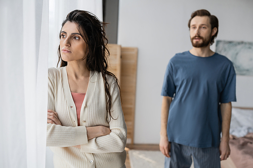 Offended woman standing near curtain while blurred boyfriend talking at home