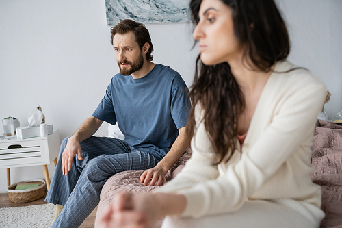 Displeased man in pajama talking to blurred girlfriend while sitting on bed