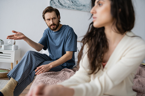 Offended man in pajama talking to blurred girlfriend while sitting on bed at home