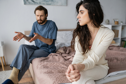 Sad woman sitting near blurred angry boyfriend talking in bedroom