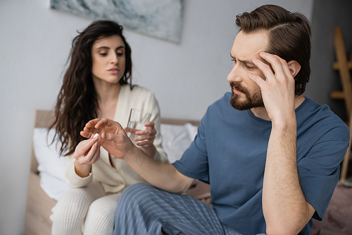 Man with headache taking pill from blurred caring girlfriend in bedroom