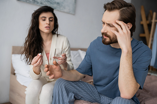 Bearded man with migraine sitting near blurred girlfriend with pills and water at home