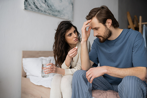 Caring woman holding pill and glass of water near boyfriend suffering from migraine in bedroom