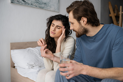 Caring man giving pills and water to girlfriend with migraine in bedroom