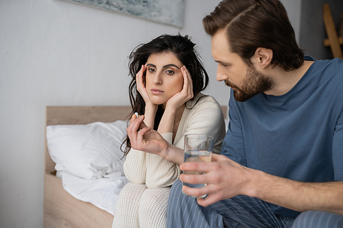 Caring man holding pills and water near frustrated girlfriend on bed at home