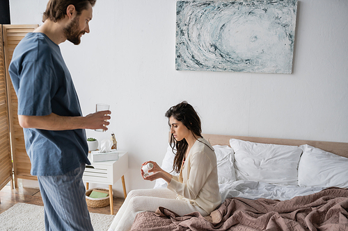 Side view of woman holding pills near caring boyfriend with glass of water in bedroom