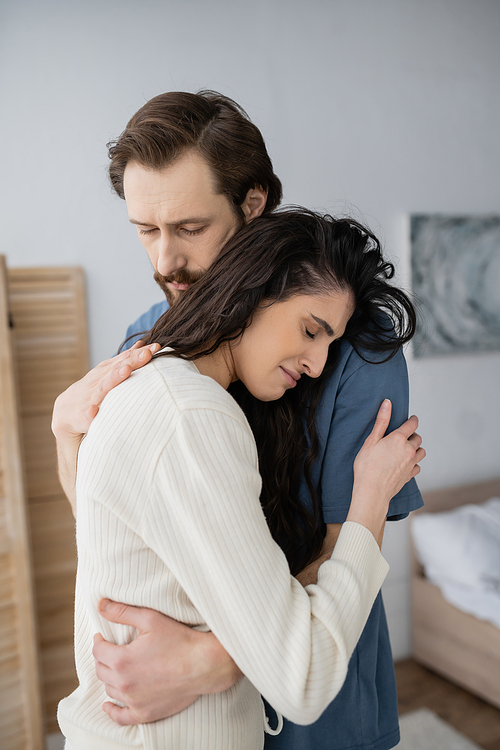 Bearded man hugging and calming displeased girlfriend in bedroom at home