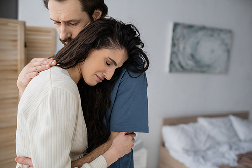 Caring man hugging crying girlfriend in blurred bedroom at home