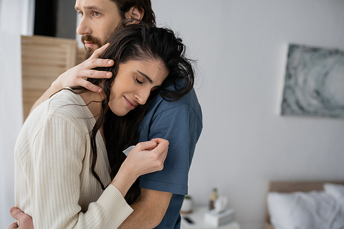 Empathic man calming crying girlfriend in blurred bedroom at home