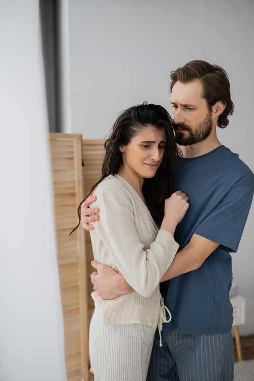 Bearded man in pajama hugging dissatisfied girlfriend at home