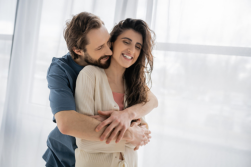 Bearded man embracing cheerful brunette girlfriend at home