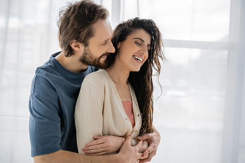 Bearded man embracing overjoyed brunette girlfriend at home