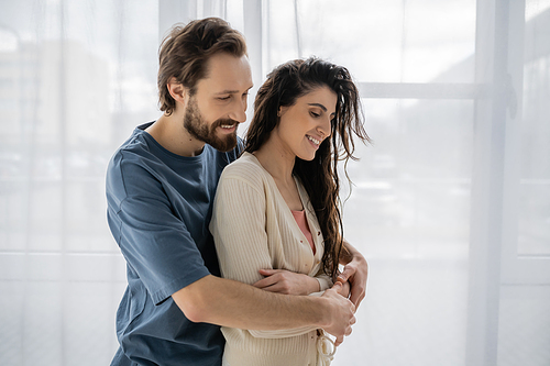 Smiling man hugging brunette girlfriend in casual clothes at home