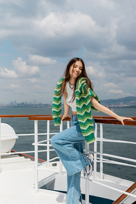 Positive young woman in knitted sweater and jeans standing near railing of yacht in Turkey