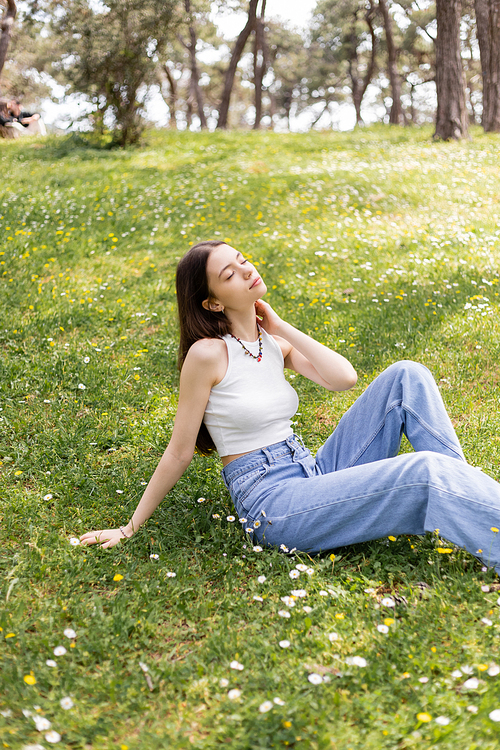 Young woman in top and jeans touching neck while sitting on lawn with flowers in park
