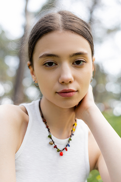 Portrait of young brunette woman in top and necklace looking at camera outdoors