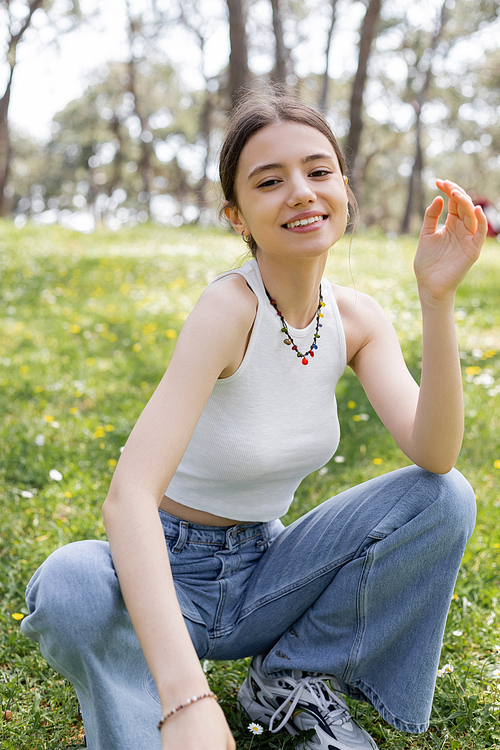 Smiling young woman in top and jeans looking at camera on blurred meadow with flowers in park