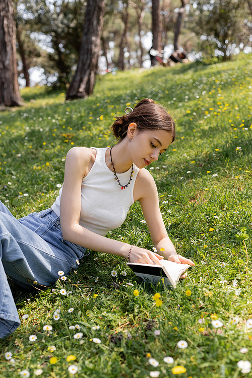 Young woman in top reading book while lying on lawn with flowers in park